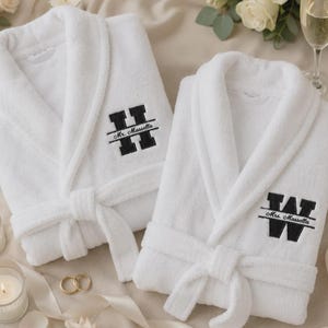 May include: Two white plush bathrobes featuring black embroidered monograms. The left robe displays a large letter H with the text Mr. Massella, while the right robe features a large letter W with the text Mrs. Massella, both with matching belts.