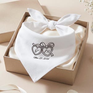 May include: White bandana with a bow, inside a gift box. The bandana features embroidered hearts with the letters "T" and "M" and the date "Mar 25, 2026". The box is beige and lined with satin.