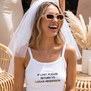 Könnte beinhalten: Ein weißes Tanktop mit dem Text "IF LOST, PLEASE RETURN TO LOGAN HENDERSON". Die Person trägt einen weißen Schleier und beige Sonnenbrille. Der Hintergrund umfasst andere Personen und dekorative Pflanzen.