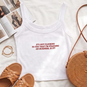 May include: White tank top with the text "IT'S NOT CLOCKING TO YOU THAT I'M STANDING ON BUSINESS, IS IT?" in red. The top is paired with brown sandals, a woven purse, and a gold bracelet. A magazine is open in the background.