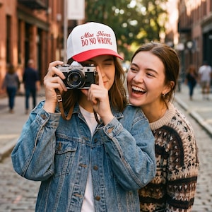 May include: A white and red baseball cap with the text "Miss Never DO NO WRONG" is worn by a person holding a Canon camera. The person is wearing a blue denim jacket. Another person is smiling next to them.