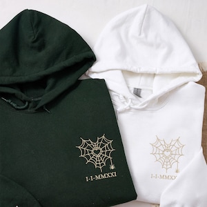 May include: Two hooded sweatshirts, one dark green and one white. Each has a gold spiderweb design with a heart and the Roman numeral date "1-I-MMXXI" or "1-I-MMXX". The sweatshirts have drawstrings and a soft texture.