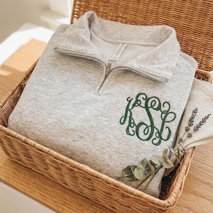 May include: A light gray pullover sweatshirt with a quarter-zip neckline and a green embroidered monogram. The sweatshirt is folded inside a woven basket, with a folded white cloth and sprigs of lavender and eucalyptus.