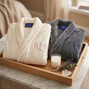 May include: Two plush bathrobes, one cream and one gray, are displayed on a wooden tray. The cream robe has embroidered initials "RLB" in pink, while the gray robe features "TAB" in blue. A candle and lavender sprigs complete the scene.