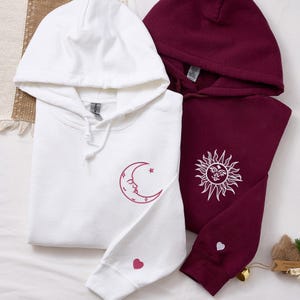 Puede incluir: Dos sudaderas con capucha, una blanca y otra burdeos. La sudadera blanca presenta un diseño de luna creciente y estrella rosa en la parte delantera y un corazón rosa en la manga. La sudadera burdeos tiene un diseño de sol blanco y un corazón blanco en la manga.