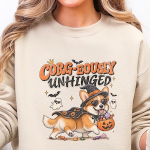 May include: A sand-colored crewneck sweatshirt featuring a graphic of a Corgi wearing a witch hat, heart-shaped sunglasses, and a pumpkin-patterned cape. The dog holds a jack-o'-lantern bucket. Text reads: CORG-EOUSLY UNHINGED.