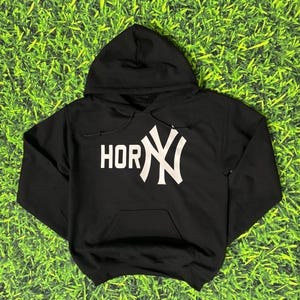 May include: Black hooded sweatshirt with a white graphic that reads "HORNY" with the New York Yankees logo. The sweatshirt has a front pocket and a drawstring hood. The background is green.