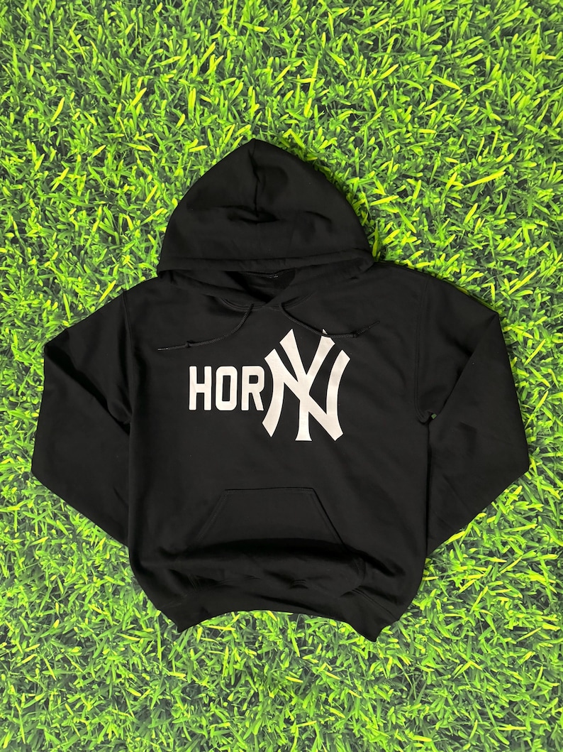 May include: Black hooded sweatshirt with a white graphic that reads "HORNY" with the New York Yankees logo. The sweatshirt has a front pocket and a drawstring hood. The background is green.