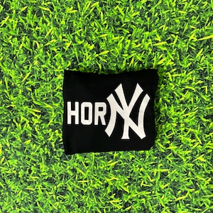 May include: Black t-shirt with the word "HOR" in white block letters and a white stylized "NY" logo. The shirt is displayed on a green grass-like background.