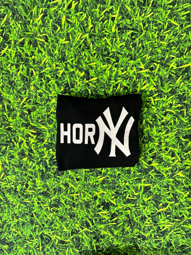 May include: Black t-shirt with the word "HOR" in white block letters and a white stylized "NY" logo. The shirt is displayed on a green grass-like background.