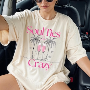 May include: Cream-colored t-shirt with the text "Soul Ties Is Crazy" in pink. The graphic includes palm trees, champagne flutes, and hearts. The shirt is a casual, oversized style.
