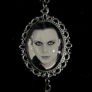 May include: Silver-toned pendant necklace featuring an oval portrait of a person with dark hair and eyes. The portrait is framed by an ornate, filigree-style border. The necklace chain is also silver-toned, with a simple link design.