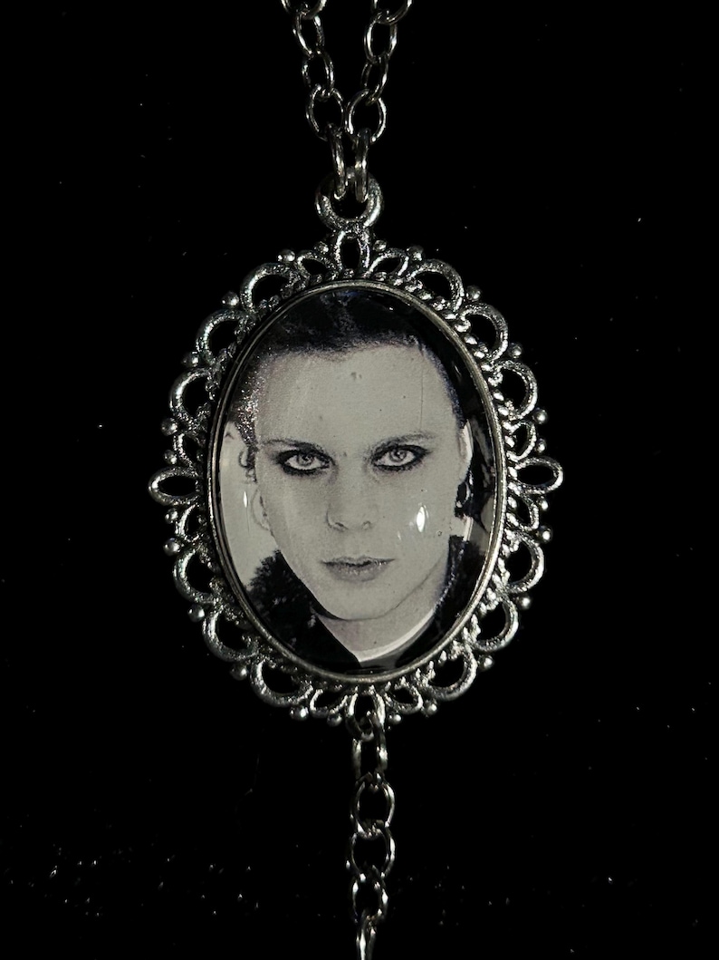 May include: Silver-toned pendant necklace featuring an oval portrait of a person with dark hair and eyes. The portrait is framed by an ornate, filigree-style border. The necklace chain is also silver-toned, with a simple link design.