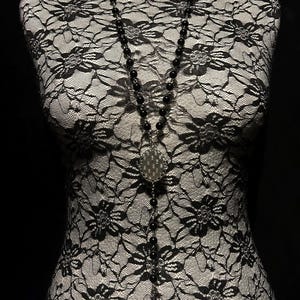 May include: A long black beaded necklace with a silver pendant featuring a dotted design. The necklace hangs on a black and white lace bust form against a black background. The beads appear to be approximately 0.25 inches in diameter.