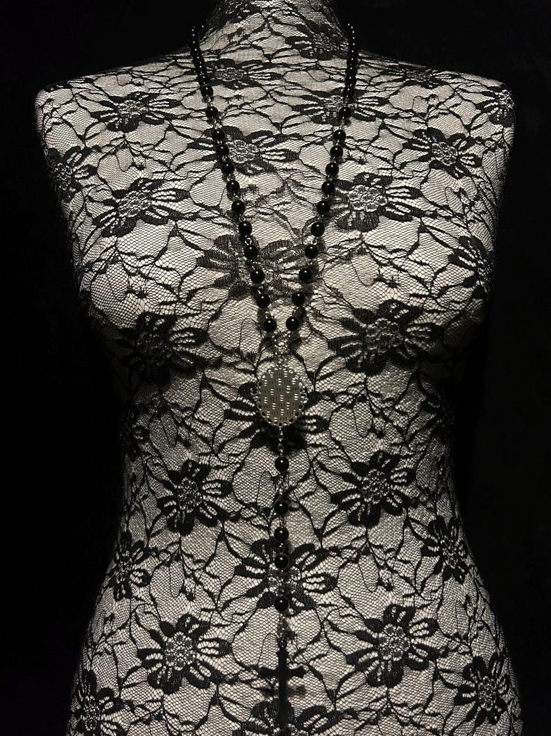 May include: A long black beaded necklace with a silver pendant featuring a dotted design. The necklace hangs on a black and white lace bust form against a black background. The beads appear to be approximately 0.25 inches in diameter.