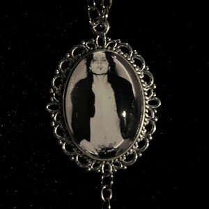 May include: Silver-toned pendant necklace featuring an oval black and white image of a person in a decorative frame. The frame has an ornate, openwork design. The necklace chain is also silver-toned and hangs against a dark background.