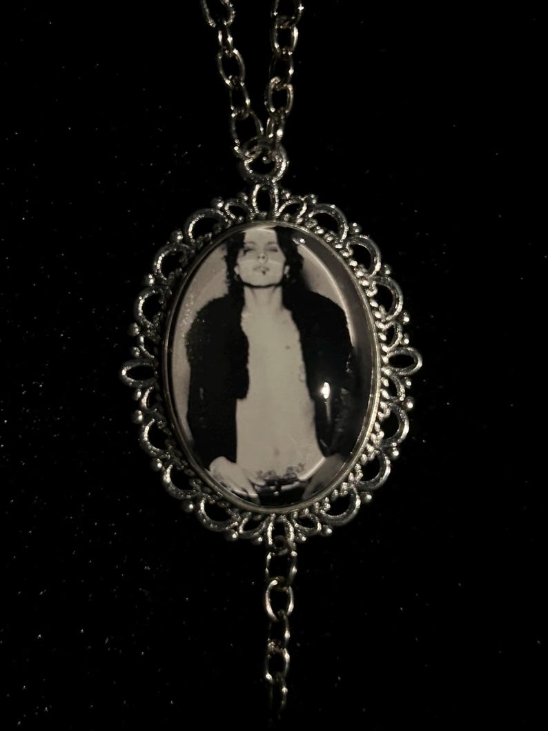 May include: Silver-toned pendant necklace featuring an oval black and white image of a person in a decorative frame. The frame has an ornate, openwork design. The necklace chain is also silver-toned and hangs against a dark background.