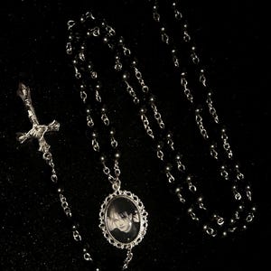 May include: A black rosary necklace with a silver cross pendant and a portrait pendant. The necklace has black beads and silver chain links. The portrait is in black and white.