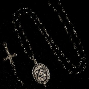 May include: A black rosary necklace with a silver-toned cross pendant and a decorative oval pendant. The oval pendant features a black heart-shaped design on a white background, framed by an ornate silver border. The beads are black.