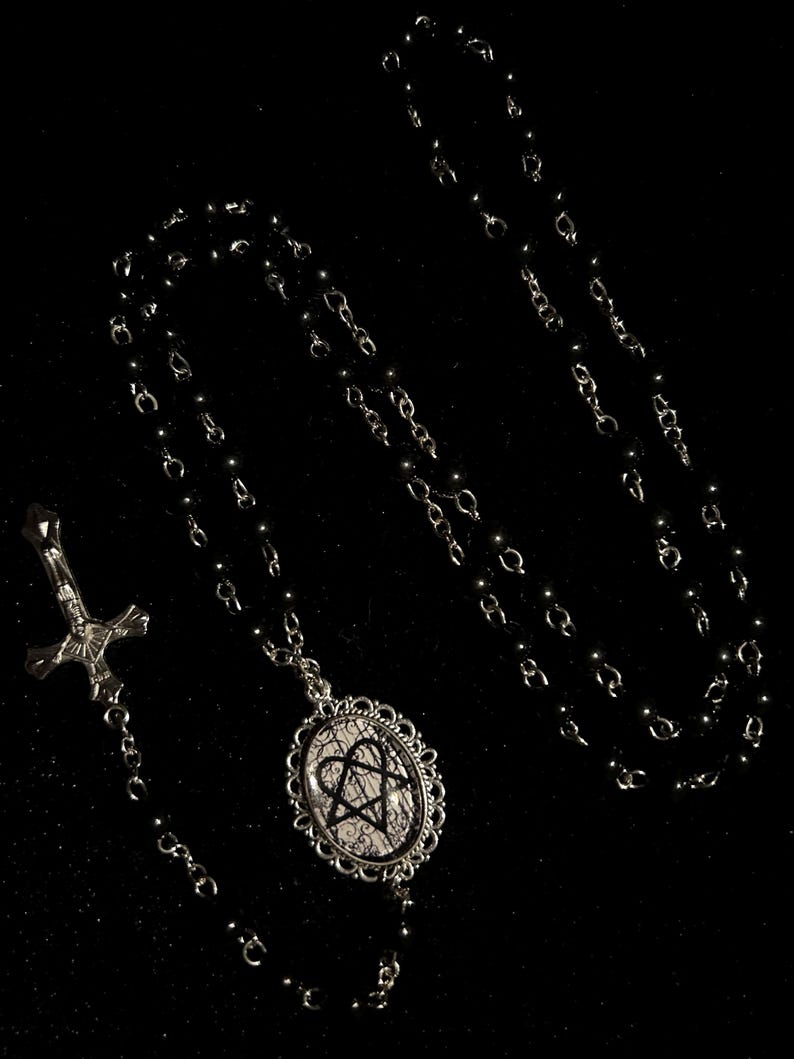 May include: A black rosary necklace with a silver-toned cross pendant and a decorative oval pendant. The oval pendant features a black heart-shaped design on a white background, framed by an ornate silver border. The beads are black.
