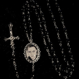 May include: A black beaded necklace with a silver-toned cross pendant and a silver-framed oval pendant featuring a black and white portrait. The necklace is displayed against a black background.