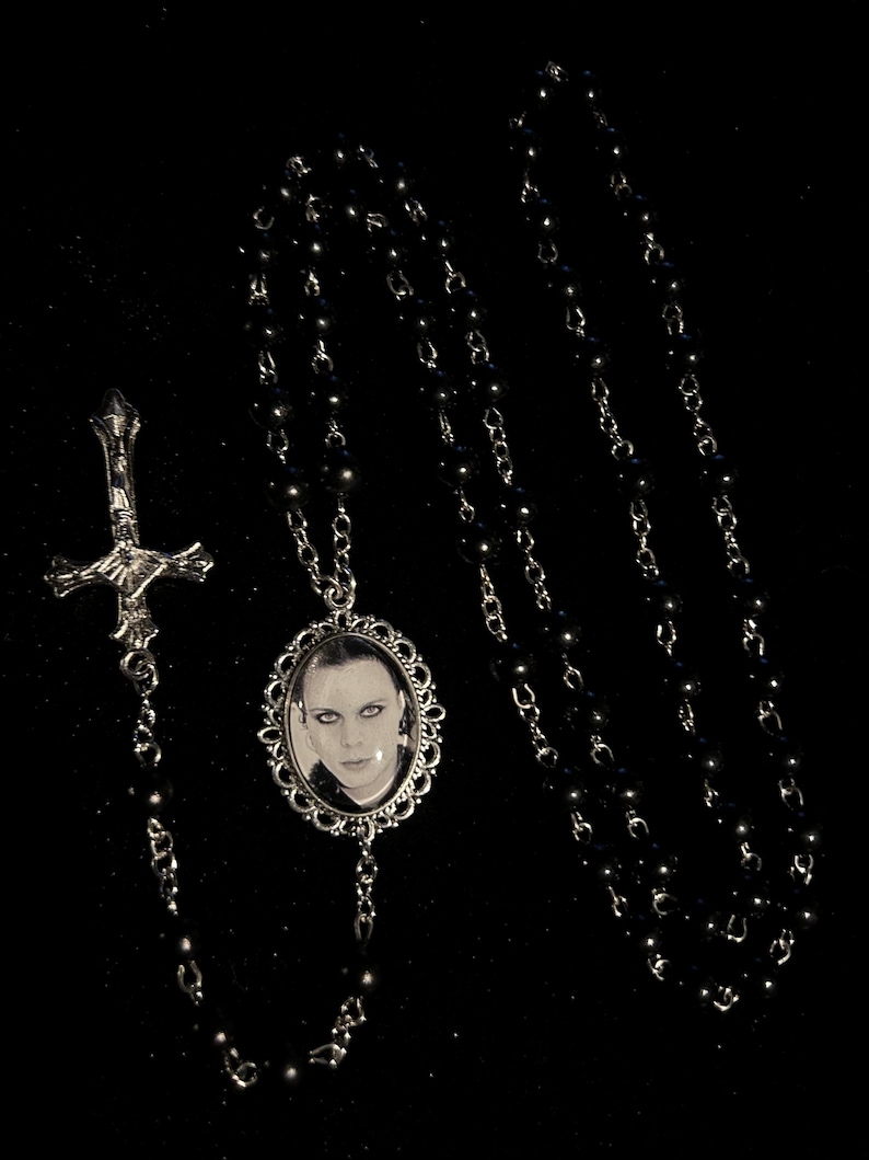 May include: A black beaded necklace with a silver-toned cross pendant and a silver-framed oval pendant featuring a black and white portrait. The necklace is displayed against a black background.