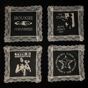 Peut inclure: Quatre dessous de verre carrés noirs avec une bordure en dentelle blanche. Chaque dessous de verre présente le nom et l'image d'un groupe : Siouxsie and the Banshees, The Cure, Type O Negative, et The Sisters of Mercy. Le texte "Boys Don't Cry" est également visible.