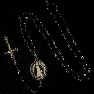 May include: A black beaded necklace with a silver pendant featuring a black and white image. The necklace includes a silver cross pendant. The jewelry is displayed against a black background.