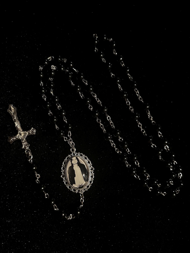 May include: A black beaded necklace with a silver pendant featuring a black and white image. The necklace includes a silver cross pendant. The jewelry is displayed against a black background.
