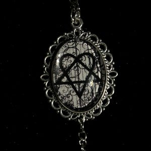 May include: Silver-toned pendant necklace with an oval-shaped charm. The charm features a black pentagram and heart symbol on a white and black patterned background. The pendant has a decorative, ornate frame and a delicate chain.