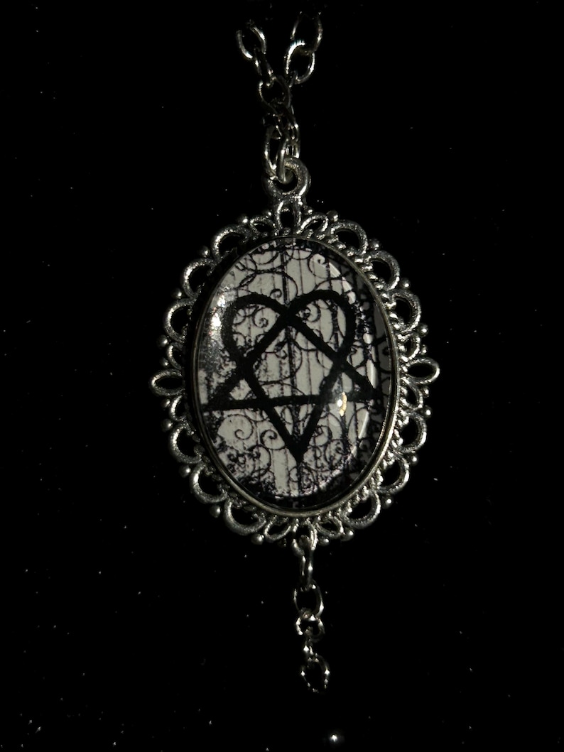 May include: Silver-toned pendant necklace with an oval-shaped charm. The charm features a black pentagram and heart symbol on a white and black patterned background. The pendant has a decorative, ornate frame and a delicate chain.