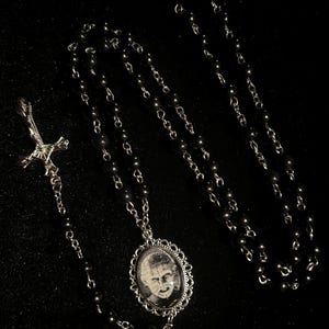 Puede incluir: Collar plateado con un colgante ovalado que muestra una imagen en blanco y negro de un rostro. El colgante tiene un borde decorativo. La cadena es de cuentas y tiene un dije de cruz.