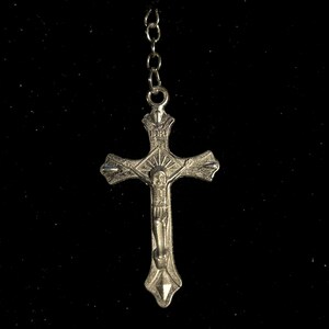 May include: A silver-colored rosary with a crucifix pendant. The crucifix features a detailed depiction of Jesus Christ. The rosary chain is connected to a series of small, dark beads. The background is black.