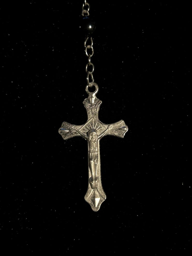 May include: A silver-colored rosary with a crucifix pendant. The crucifix features a detailed depiction of Jesus Christ. The rosary chain is connected to a series of small, dark beads. The background is black.