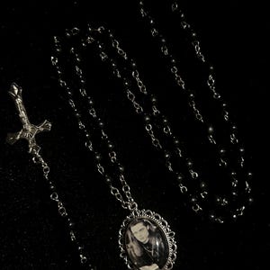 May include: A silver necklace with a pendant displaying a black and white portrait of a person with long hair. The necklace features a chain of small, dark beads and a cross pendant.