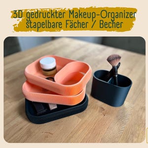 Könnte beinhalten: Stapelbarer Make-up-Organizer in Koralle und Schwarz. Der Organizer enthält Tabletts und einen geteilten Behälter für Pinsel. Der Text auf dem Bild lautet "3D gedruckter Makeup-Organizer stapelbare Fächer / Becher."