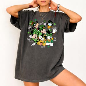Puede incluir: Camiseta gris oscuro con un diseño del Día de San Patricio. Presenta a los personajes de dibujos animados Mickey Mouse, Minnie Mouse, Goofy, Donald Duck, Daisy Duck y Pluto, todos con trajes y sombreros verdes, con tréboles.