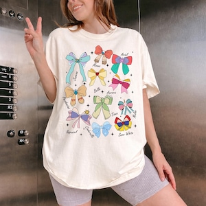 May include: A cream-coloured t-shirt displaying colourful bow illustrations with character names such as Ariel, Belle, and Snow White. The bows are in various colours and styles, with names written in a script font.