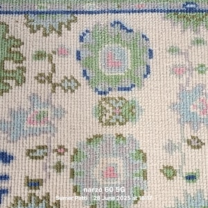 Op de afbeelding: Een geweven tapijt met een gebroken witte basis en een herhalend patroon van gestileerde bloem- en geometrische vormen in tinten saliegroen, lichtblauw, donkerblauw en zachtroze. Het tapijt heeft een blauwe rand en bevat de tekst narzo 60 5G.