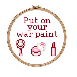 May include: Embroidered hoop art with the text "Put on your war paint" in red. The hoop features a pink lipstick, a pink compact with a white bow, and a pink hand mirror.