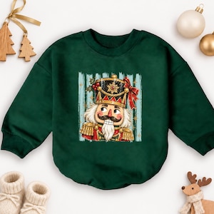 May include: A dark green long-sleeve sweatshirt featuring a central graphic print of a nutcracker soldier. The nutcracker wears a black and gold hat with a red bow, a red uniform with gold epaulets, and has a white mustache and beard.