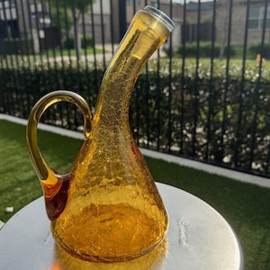 May include: A decorative, amber-colored glass pitcher with a curved neck and a handle. The pitcher has a cracked, textured surface and sits on a silver metal surface. The pitcher is likely used for serving beverages.