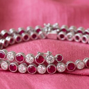 May include: A silver bracelet with two rows of alternating red and clear gemstones. The bracelet is set against a pink fabric background, creating a contrast that highlights the jewelry's sparkle and design.