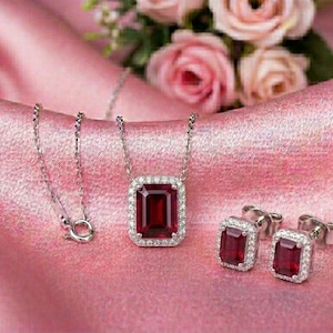 May include: A silver necklace with a rectangular, deep red gemstone pendant, surrounded by small white stones. Matching stud earrings with similar red gemstones and white stone borders are also present. The jewelry rests on a pink satin surface, with pink roses in the background.