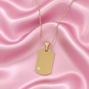 May include: A gold dog tag pendant with a small diamond, hanging from a fine gold chain. The pendant is rectangular with rounded corners. The background is a pink satin fabric.