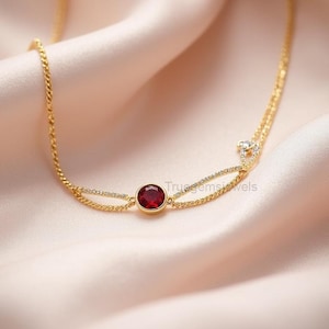 May include: A delicate gold necklace with a double chain design. The necklace features a round, deep red gemstone as a focal point, complemented by small, sparkling accents. The jewelry rests on a soft, peach-colored fabric.