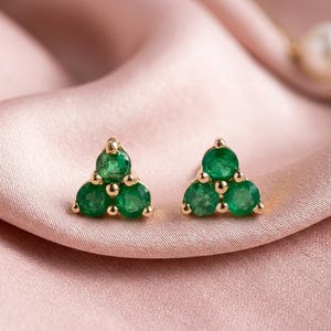 May include: A pair of gold stud earrings, each featuring three vibrant green emeralds arranged in a triangular cluster. The earrings are set against a soft, pink background, highlighting their color and design.