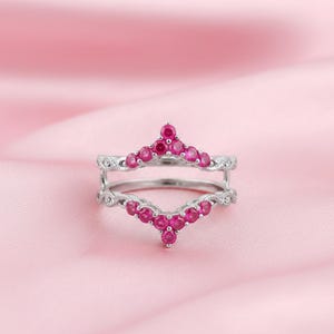 May include: A silver ring with a V-shaped design, adorned with numerous small, round, bright pink gemstones. The ring has a double band and is set against a soft pink background.
