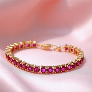 May include: A gold-toned bracelet with a row of vibrant, square-cut, pink gemstones. The bracelet is displayed on a soft, pink fabric, creating a luxurious and elegant presentation. The clasp is visible, and the overall design is symmetrical.