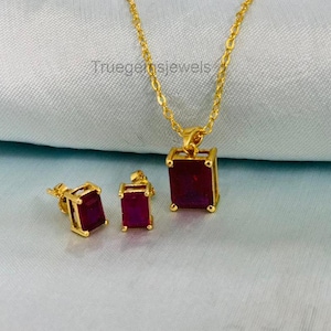 May include: A gold-colored jewelry set featuring a necklace with a pendant and matching stud earrings. Each piece showcases a rectangular, deep red gemstone. The text "Truegemsjewels" is visible on the surface.